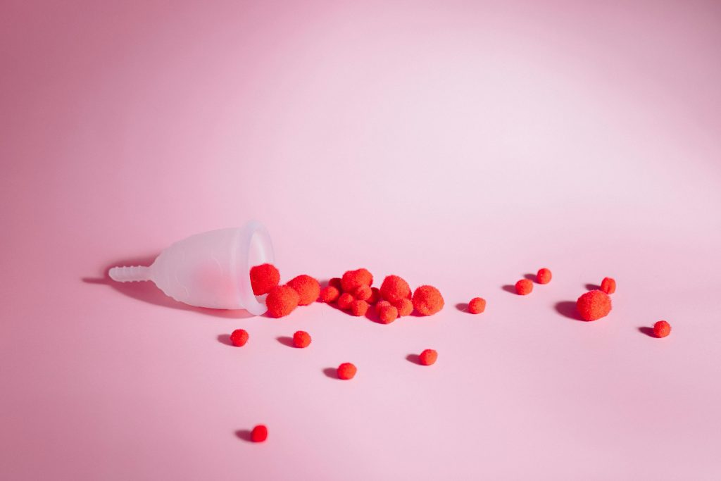 pexels-photo-5712298-5712298 Menstrual cup with red pom poms on pink background symbolizing menstruation and feminine hygiene.